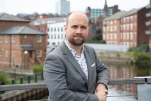 Photo of a white man in a jacket standing in front of a city scape