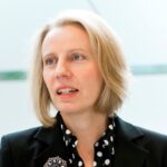 A white woman with blond hair wearing a black jacket and black blouse with white spots
