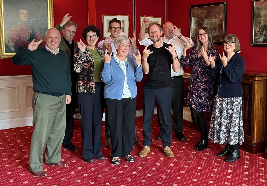 Group of 9 adults in business wear stand in a room with red carpet and wall paper, making a symbol of their hands associated with heave metal music.