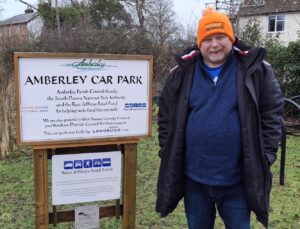 A man with a short beard wearing an orange bobble hat, blue jumper and anorak stands outside next to a sign which refers to Amberley Car Park and funding from Rees Jeffreys Road Fund