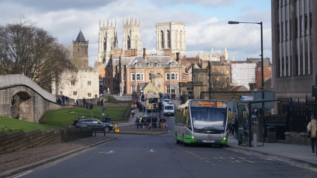 An image of a road junction in York, with a bus, cars and pedestrians in front of historic buildings.