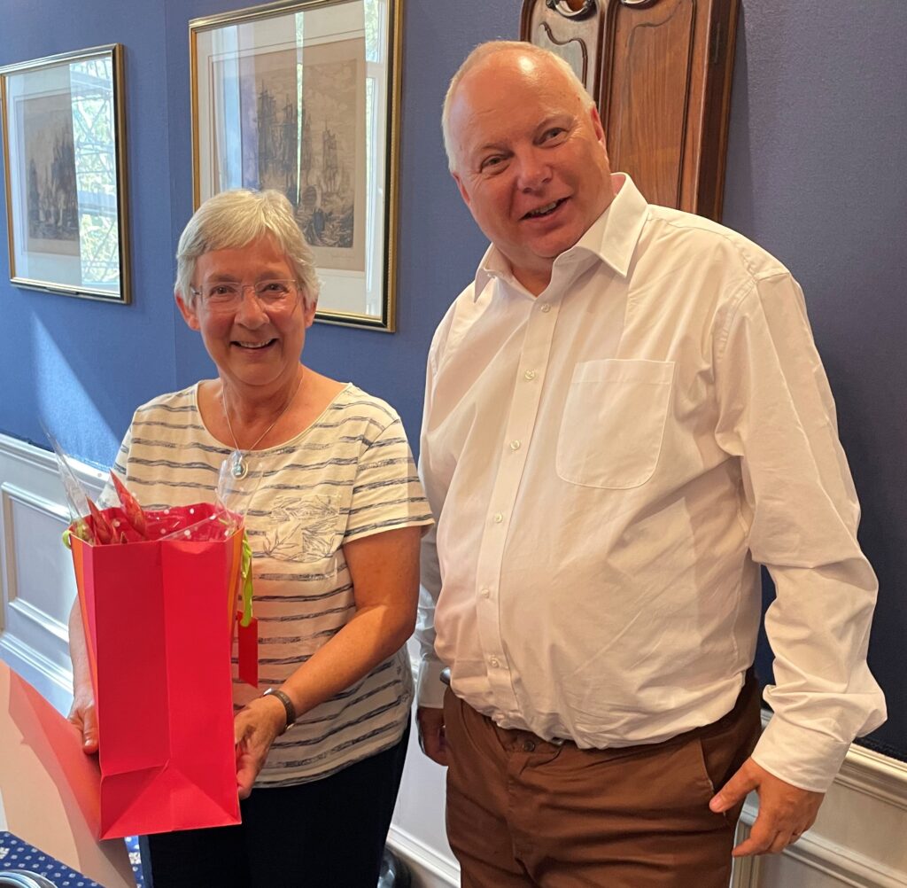 A lady in a white tshirt holds a red gift bag standing next to a taller man in a white shirt. they are both smiling.