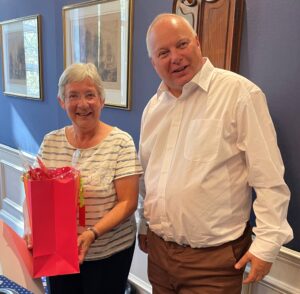 A lady in a white tshirt holds a red gift bag standing next to a taller man in a white shirt. they are both smiling.