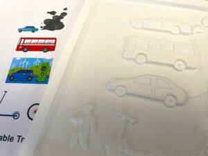 There is a partial view of a page of a 3D book and a partial view of a page of a printed colour paper-based book. They show the same images - side-on profile of a saloon car emitting clouds of exhaust fumes; a bus; a saloon car connected to an electric vehicle charging point by a lead; a scooter. The 3 D book is white. the paper based book has some colour - the bus is red; the cars are blue.
