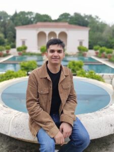 A young male in a black t-shirt and brown jacket sits in front of an Italian-style garden