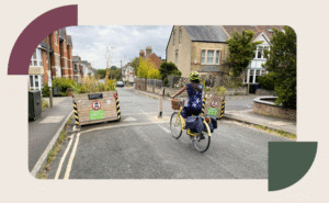 A female in a blue flowery t-shirt sits on a yellow bicycle, cycling down a tarmac residential road. The road is narrowed by two wooden planters with signs indicating the road ahead is closed to cars and motorcycles.