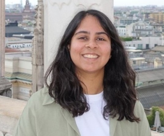 Photo of a young female with long dark hair white top and green shirt, smiling.