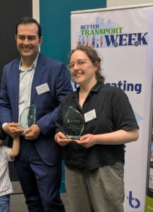 Future Transport Visions Group 2025 winners Ruth Carlson and Nima Dadash-Zadeh - a woman wearing glasses, a black top and grey trousers holds a glass plaque, standing next to a man wearing a grey suit and white shirt holding a similar glass plaque