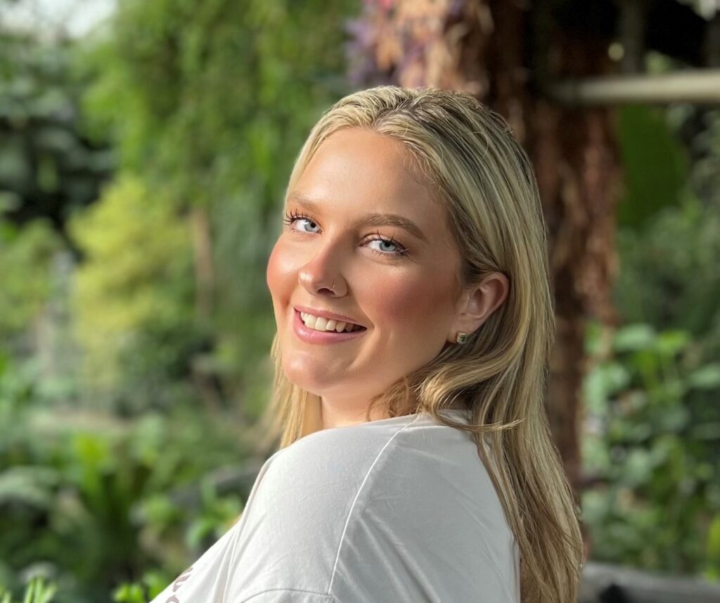 A young female with long dark straight blond hair wearing a white t-shirt and standing in front of some greenery is smiling at the camera.