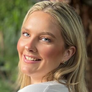 A young female with long dark straight blond hair wearing a white t-shirt and standing in front of some greenery is smiling at the camera.