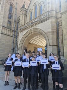 13 school children in dark school uniform with white shirts stand in front of an arched entrance doorway saying University of Manchester. They are holding up certificates.