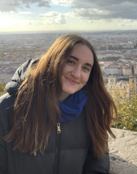 Photo of a young woman with long dark straight hair, wearing a dark coat and blue scarf standing in an elevated position with a townscape behind her in the distance. She is smiling at the camera.