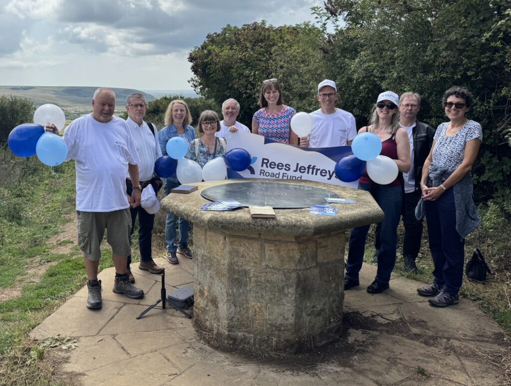 10 people stand behind a large stone monument in the shape of an octagonal table. There are trees and a view of the coast in the background. They are holding balloons.