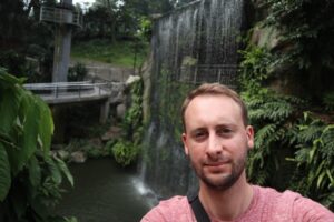 A fair haired man with a beard wearing a pink t-shirt is standing in front of a waterfall with a viewing platform and ferns