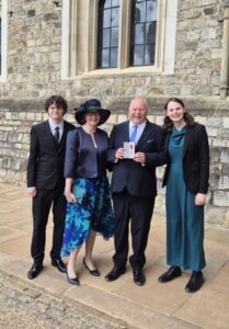 A man in a suit holds his MBE accompanied by his wife in a hat, and son and daughter.